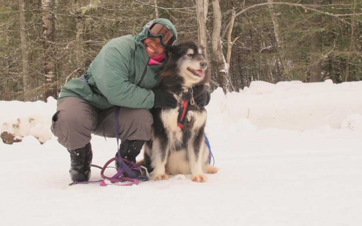 a person wearing snow gear smiles and bends down to hug a sled dog